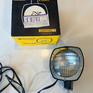May include: A Penncrest Super 8 sealed beam movie light with a black and yellow cardboard box. The light features a square black housing with a chrome face and a circular glass lens. A black power cord is attached to the base of the light unit.