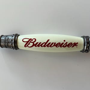 May include: A Budweiser tap handle with a cream-colored body and the Budweiser logo in red script. The handle has metal accents on each end and a metal spout. The tap handle is designed for a beer keg.