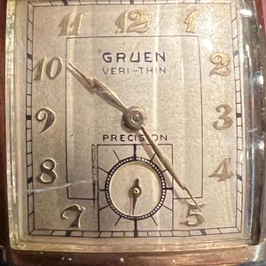 May include: Vintage Gruen Veri-Thin watch with a rectangular face. The watch has gold-toned hands and numerals against a light-colored background. The watch face is marked with the brand name and "Precision". The watch has a gold-toned, twisted rope-style band.