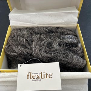 May include: A gray wig with a curly style, presented in a yellow box with tissue paper. The wig is labeled "flexlite" with "KANEKALON MODACRYLIC" below the brand name.