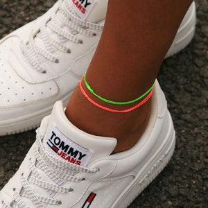 Może przedstawiać: Para białych butów sportowych z logo Tommy Jeans z boku. Osoba nosząca buty ma dwie neonowe bransoletki na kostkę, jedną zieloną i jedną różową.