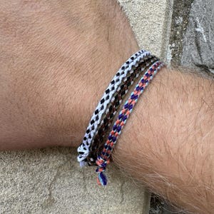 May include: Three colourful friendship bracelets stacked on a wrist. The bracelets are made of woven string in shades of white, black, brown, orange, blue, and purple.