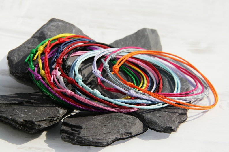 Pu&ograve; includere: Una collezione di braccialetti in corda intrecciata colorati, in vari colori, tra cui rosso, blu, verde, giallo, rosa e viola. I braccialetti sono disposti in cerchio su una superficie di pietra grigia.