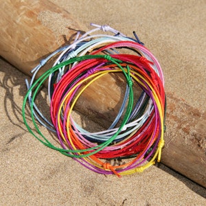 Pu&ograve; includere: Una collezione di braccialetti di corda colorati in vari colori, tra cui rosso, blu, verde, giallo e bianco. I braccialetti sono disposti in cerchio su una superficie di legno.
