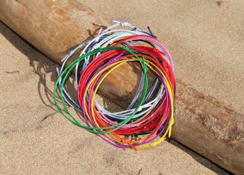 Pu&ograve; includere: Una collezione di braccialetti di corda colorati in vari colori, tra cui rosso, blu, verde, giallo e bianco. I braccialetti sono disposti in cerchio su una superficie di legno.