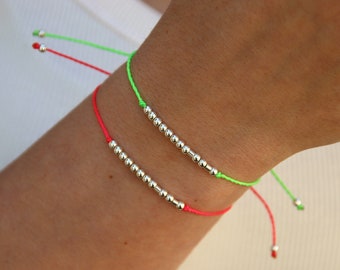 May include: Two minimalist bracelets. One is neon green, the other is neon pink. Each bracelet features a string of small, silver-colored beads. The bracelets are worn on a wrist.