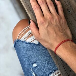 Pu&ograve; includere: Una mano con smalto nero per unghie riposa su una superficie di legno. Un braccialetto di corda rosso &egrave; indossato al polso.