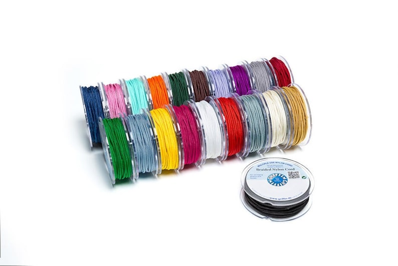 Pu&ograve; includere: Una collezione di bobine di cordoncino in nylon intrecciato in vari colori, tra cui rosso, verde, blu, giallo, arancione, bianco e nero. Le bobine sono disposte in fila, con la bobina nera in primo piano. Il testo "Braided Nylon Cord" &egrave; visibile sull'etichetta della bobina nera.
