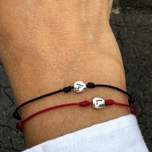 May include: Two friendship bracelets with black and red string. The bracelets have silver charms with the letters "A" and "L" on them.