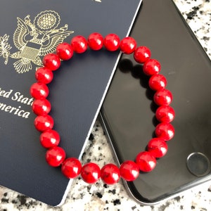 May include: A red beaded bracelet with black speckles. The bracelet is lying on top of a blue passport and a black smartphone.