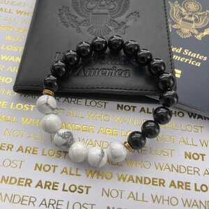 May include: A black and white beaded bracelet with gold accents. The bracelet is resting on a black leather passport holder with the words "United States of America" embossed on it. The bracelet is also resting on a blue passport with the words "United States of America" printed on it.
