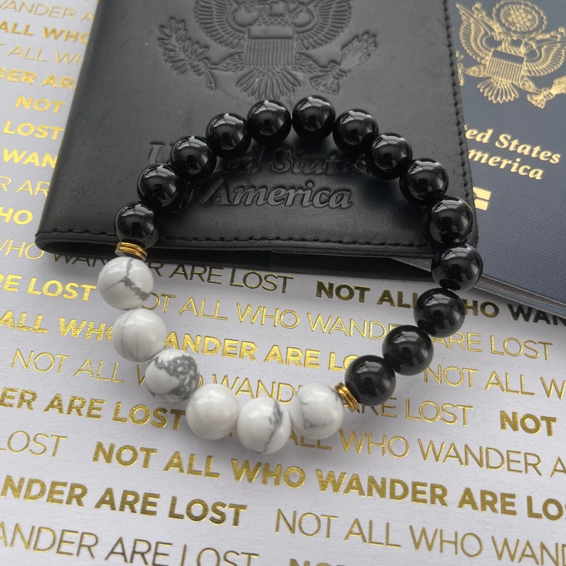 May include: A black and white beaded bracelet with gold accents. The bracelet is resting on a black leather passport holder with the words "United States of America" embossed on it. The bracelet is also resting on a blue passport with the words "United States of America" printed on it.