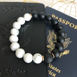 May include: A black and white beaded bracelet with a lava rock bead. The bracelet is on top of two passports, one black and one blue. The passports have the words "PASSPORT" and "United States of America" printed on them.