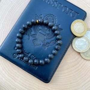 May include: A black leather passport holder with the word "PASSPORT" embossed on the cover. A black lava rock beaded bracelet with a gold accent is lying on top of the passport holder.