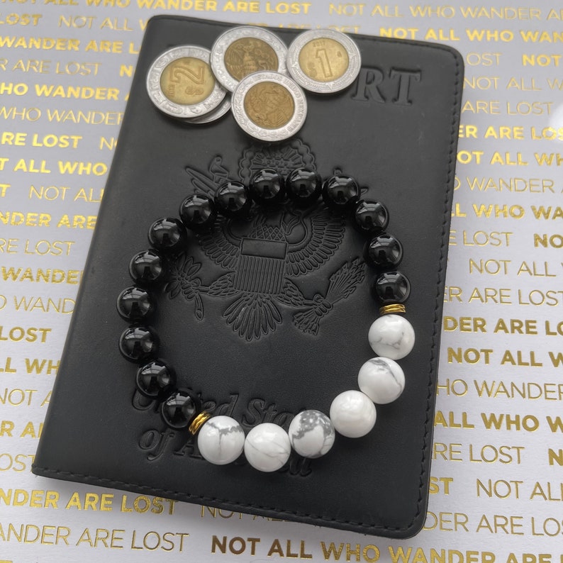 May include: A black and white beaded bracelet with gold accents. The bracelet is lying on top of a black leather passport holder with the words "Not All Who Wander Are Lost" printed in gold on the cover.