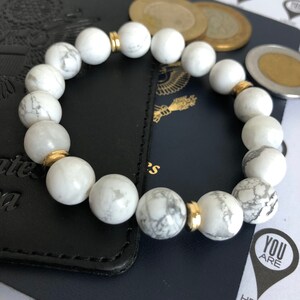 Peut inclure: Bracelet en perles d'howlite blanche avec des accents dor&eacute;s. Le bracelet est sur une surface en cuir noir.
