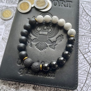 May include: A black and white beaded bracelet with gold accents. The bracelet is made with black lava rock beads, black onyx beads, and white howlite beads. The bracelet is lying on a black leather passport holder.