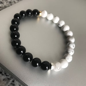 May include: A black and white beaded bracelet with a gold accent. The bracelet is made of black onyx and white howlite beads.