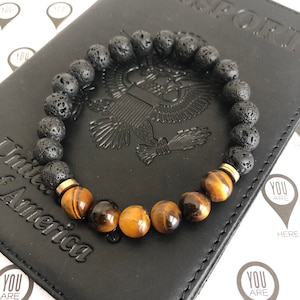 May include: A black lava rock and tiger's eye beaded bracelet with gold spacers. The bracelet is lying on a black leather surface with the words "United States of America" embossed on it.