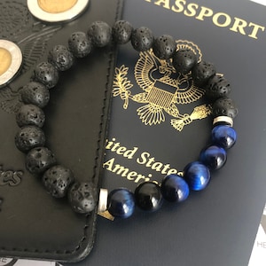 Pode incluir: Um bracelete de contas de lava preta e olho de tigre azul com detalhes prateados. O bracelete está sobre um passaporte dos Estados Unidos.