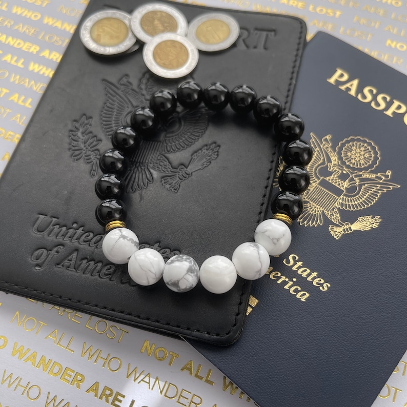 May include: A black and white beaded bracelet with gold accents. The bracelet is lying on top of a black leather passport holder and a blue passport. The passport holder has the words "United States of America" embossed on it. The passport has the words "PASSPORT" printed on it.