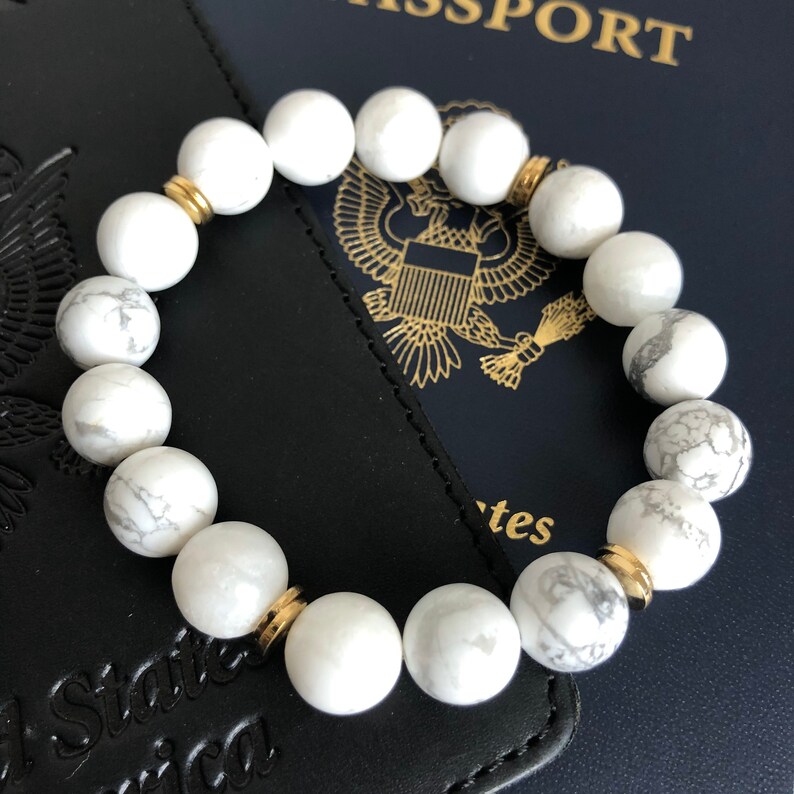Peut inclure: Bracelet en perles d'howlite blanche avec des accents dor&eacute;s. Le bracelet est pos&eacute; sur un passeport noir.