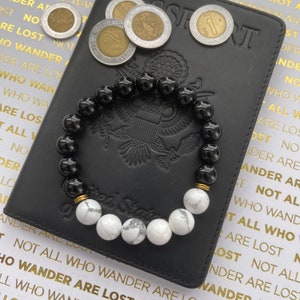 May include: A black and white beaded bracelet with gold accents. The bracelet is lying on a black leather passport with the words "United States of America" embossed on the cover.