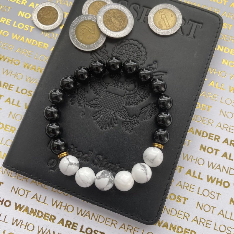 May include: A black and white beaded bracelet with gold accents. The bracelet is lying on a black leather passport with the words "United States of America" embossed on the cover.