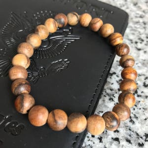 Op de afbeelding: Een kralenarmband van ronde, lichtbruine houten kralen met donkerbruine nerfpatronen. De armband ligt op een zwart leren oppervlak met reliëfdetails. De kralen hebben een diameter van ongeveer 1,3 cm.