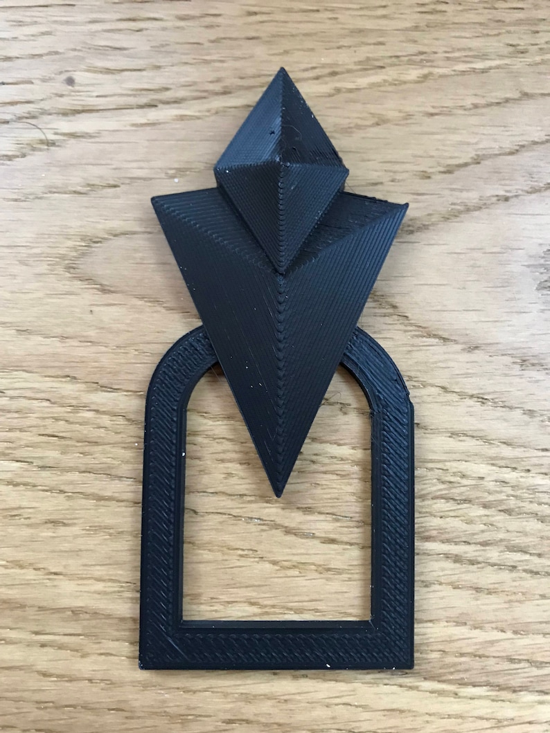 Pu&ograve; includere: Un design geometrico nero stampato in 3D con una parte superiore appuntita e una base rettangolare. Il design ricorda un arco stilizzato.