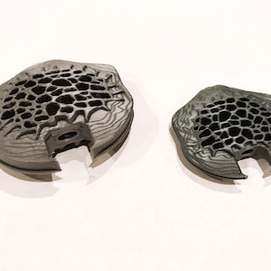 May include: Two 3D printed gray plastic objects with a honeycomb pattern. The larger object is slightly larger than the smaller object.