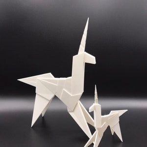 May include: Two white origami unicorns, one larger than the other, stand on a black surface. The larger unicorn has a long, pointed horn, while the smaller unicorn has a shorter horn.