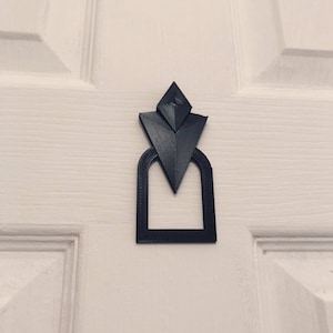 Pu&ograve; includere: Un batacchio per porta geometrico nero con un design triangolare. Il batacchio &egrave; fissato a una porta bianca.