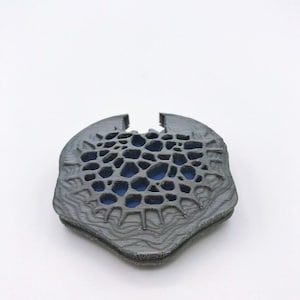 May include: A 3D printed, gray, geometric design with a blue center. The design is intricate and has a textured surface.