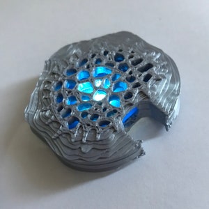 May include: A 3D printed, silver-colored object with a blue light shining through a geometric pattern.