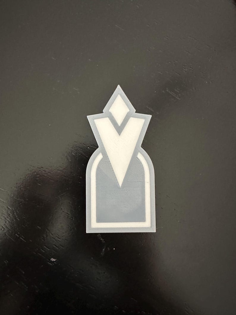 Pu&ograve; includere: Un emblema stampato in 3D grigio e bianco con una forma a V stilizzata e un diamante in cima.