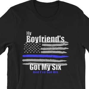 Può includere: Maglietta nera con una bandiera americana blu e bianca con una linea blu al centro. Il testo "My Boyfriend's Got My Six And I've Got His" è stampato sotto la bandiera.
