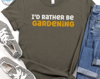 Può includere: Una maglietta verde con il testo "I'd Rather Be Gardening" in lettere bianche e arancioni. La maglietta è stesa su una superficie bianca con un paio di scarpe a righe e un paio di jeans blu.
