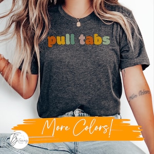 Op de afbeelding: Een grijs T-shirt met de tekst "pull tabs" in kleurrijke letters. De tekst is in een retro-lettertype. Het shirt wordt gedragen door een persoon. De tekst "More Colors!" is in wit op een gele achtergrond geschreven.
