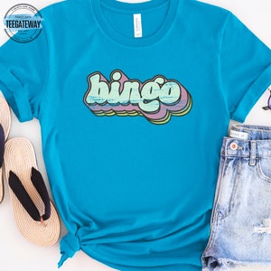 May include: A turquoise blue t-shirt with the word "bingo" printed in a retro style with a rainbow color scheme.