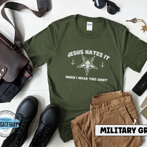 May include: A green t-shirt with the text "JESUS HATES IT" and a pentagram with a goat's head inside. The text "WHEN I WEAR THIS SHIRT" is below the pentagram. The t-shirt is on a white wooden surface with a brown leather bag, a pair of sunglasses, a pair of brown pants, and a pair of black boots.