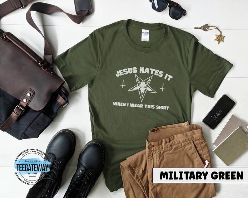 May include: A green t-shirt with the text "JESUS HATES IT" and a pentagram with a goat's head inside. The text "WHEN I WEAR THIS SHIRT" is below the pentagram. The t-shirt is on a white wooden surface with a brown leather bag, a pair of sunglasses, a pair of brown pants, and a pair of black boots.