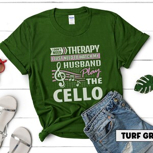 Pu&ograve; includere: Una maglietta verde con il testo "I don't need therapy I just need to watch my husband play the cello" in bianco e rosa. Il testo &egrave; circondato da note musicali. La maglietta &egrave; su una superficie bianca con un paio di jeans e una foglia verde.