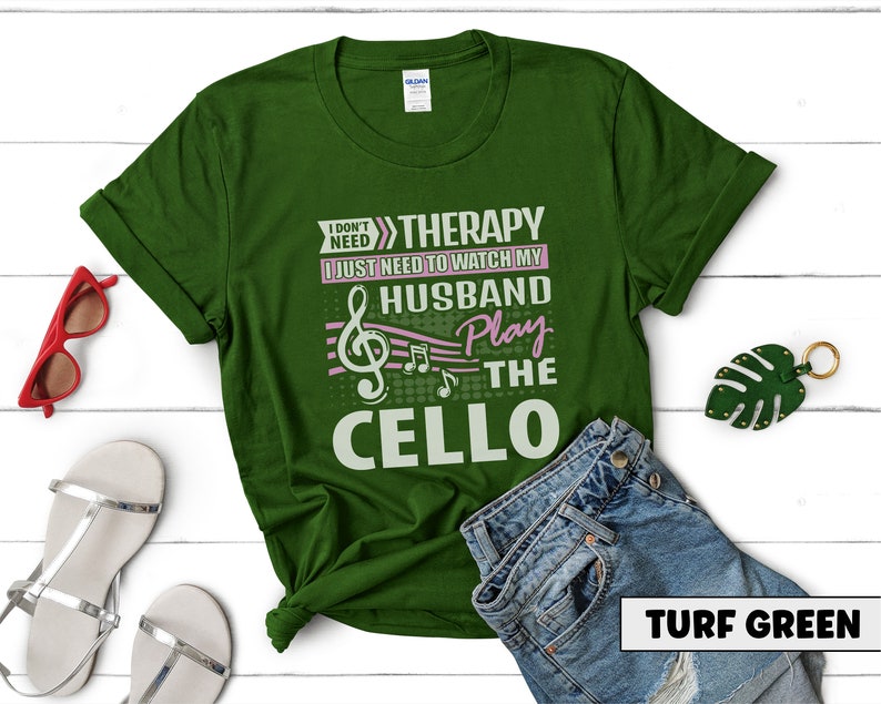 Pu&ograve; includere: Una maglietta verde con il testo "I don't need therapy I just need to watch my husband play the cello" in bianco e rosa. Il testo &egrave; circondato da note musicali. La maglietta &egrave; su una superficie bianca con un paio di jeans e una foglia verde.