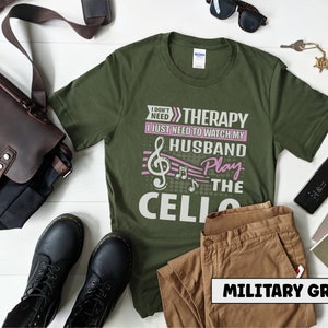 Pu&ograve; includere: Una maglietta verde con il testo "I don't need therapy I just need to watch my husband play the cello" in lettere bianche e rosa. Il testo &egrave; accompagnato da una nota musicale e note musicali.