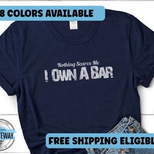 Pode incluir: Uma camiseta azul marinho com texto branco que diz "Nothing Scares Me I Own A Bar". A camiseta está disponível em 8 cores e oferece frete grátis.