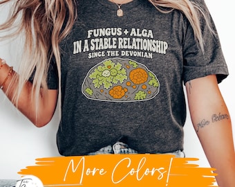 Può includere: Una t-shirt grigio scuro mélange con la grafica di una roccia con licheni verdi e arancioni. Il testo recita: FUNGUS + ALGA IN A STABLE RELATIONSHIP SINCE THE DEVONIAN. Il disegno include due piccole faccine sorridenti sul lichene verde.