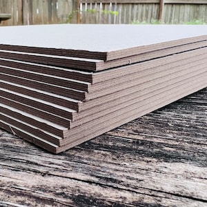May include: A stack of 10 light gray rectangular sheets of drywall, stacked on a weathered wooden surface.