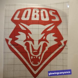Peut inclure: Autocollant en vinyle rouge avec le mot "LOBOS" et une tête de loup stylisée. L'autocollant est sur une feuille de transfert transparente avec un motif quadrillé. Le texte "glowingcanyonco" est en bas à droite.