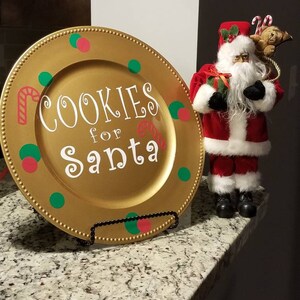 May include: A gold charger plate with red and green polka dots and the text "Cookies for Santa" in white. A Santa Claus figurine is standing to the right of the plate.